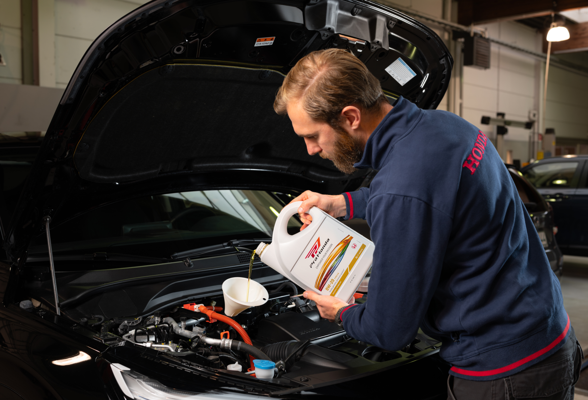 A mechanic wearing a Honda-branded uniform pours oil into a car engine from a large white container labeled \"Pro Honda\". The car's hood is open, revealing the engine compartment of what appears to be a Honda vehicle.